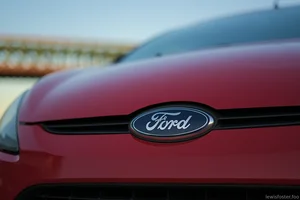 A close-up photograph of the front of a red Ford car, focusing on the central badge and grille, with a softly blurred bridge and sky in the background.