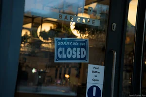 A café door at dawn displays a hanging sign reading “Sorry, we are closed,” with warm interior lights glowing inside and street reflections layered across the glass.