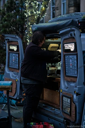 A person prepares drinks inside the open back of a mobile coffee van at sunrise, with illuminated menu boards on the open doors and soft morning light filtering through trees wrapped in string lights.